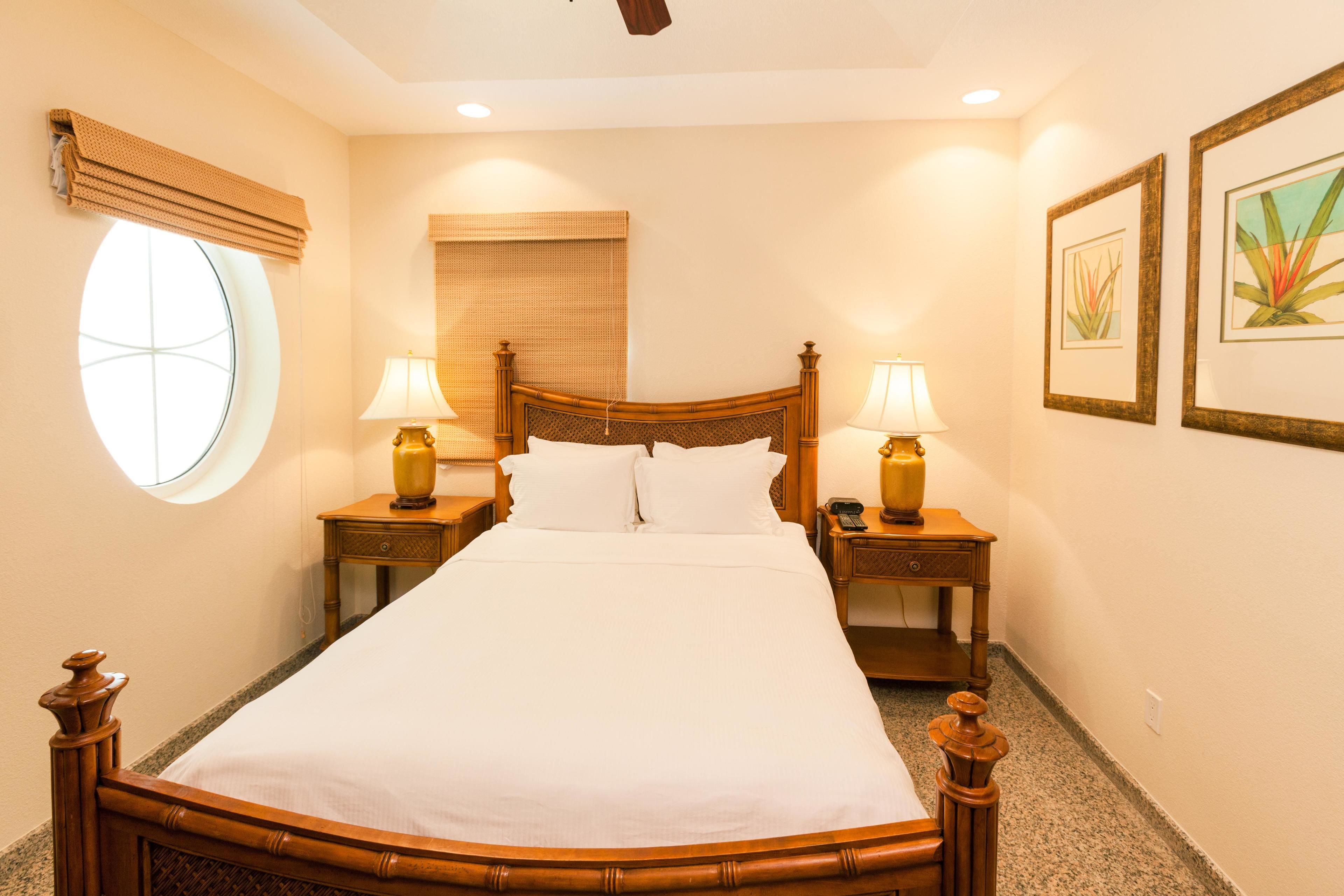 A neatly made bed with a wooden headboard, two bedside tables with lamps, a window with a blind, and two framed pictures on the wall.
