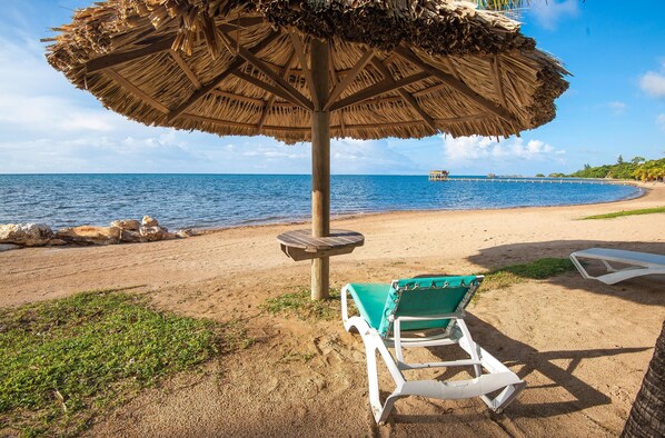 A beach with a thatched umbrella, a green lounge chair, and a view of the ocean.