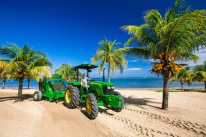 A green tractor on a sandy beach with palm trees and the ocean in the background.