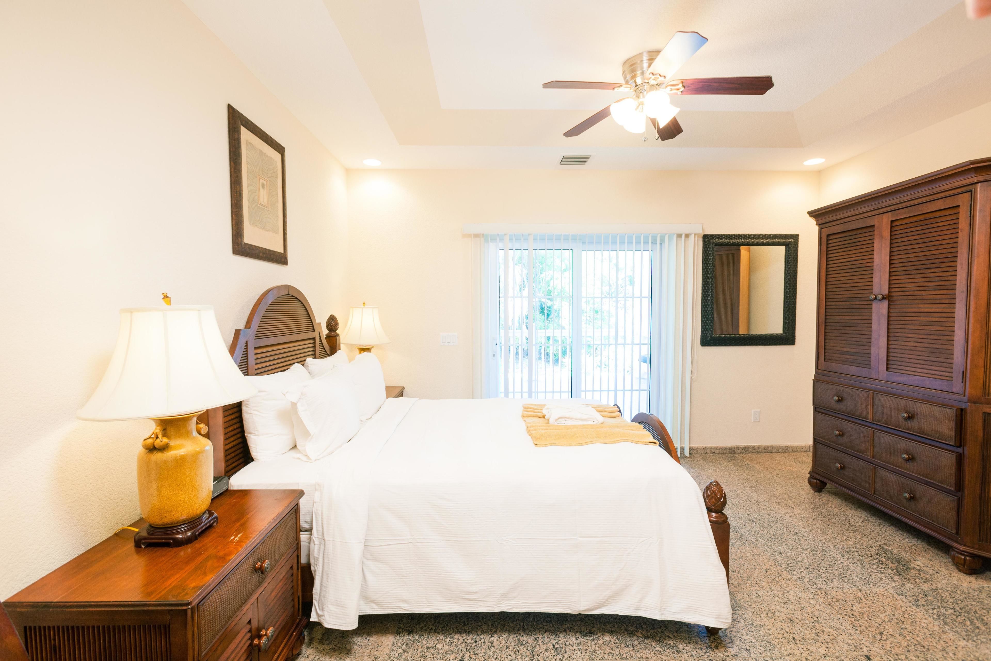 A bedroom with a bed, two bedside tables with lamps, a wardrobe, a ceiling fan, and a window with blinds.