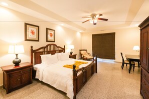 A hotel room with a wooden bed, two bedside tables with lamps, a ceiling fan, a dining table with chairs, and framed artwork on the walls.