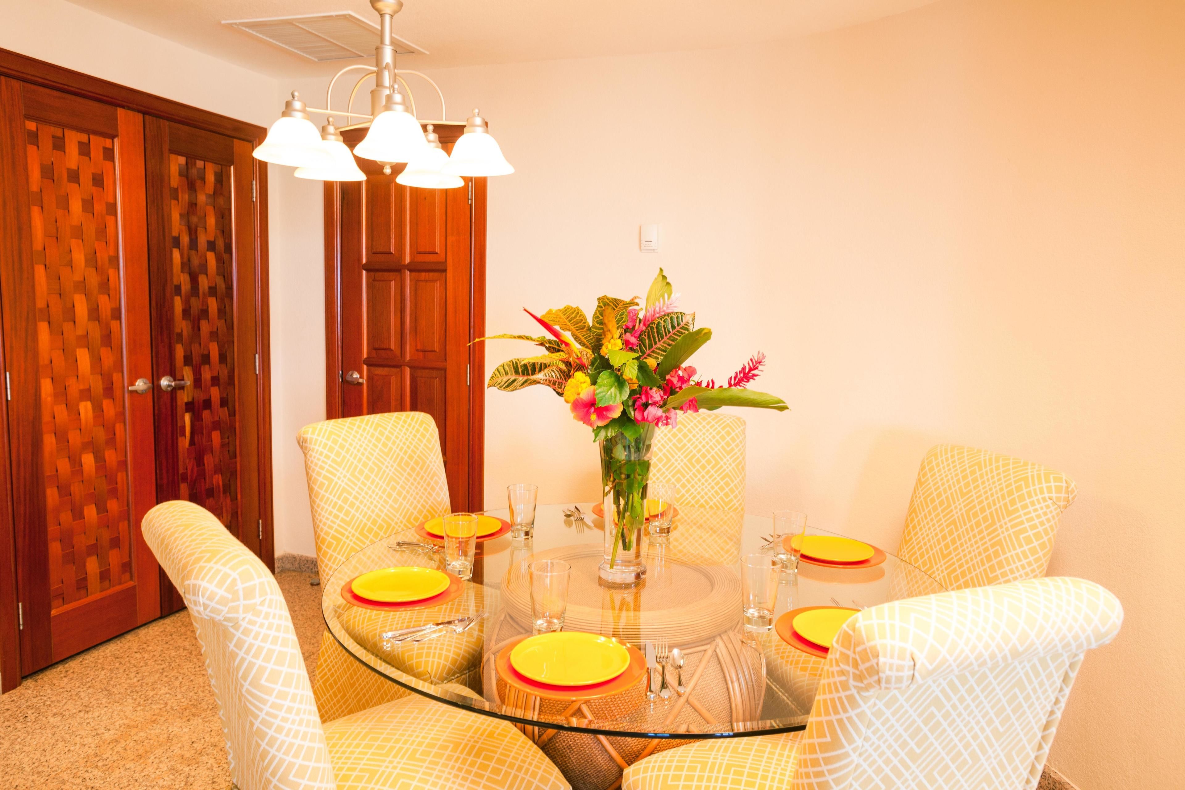 A dining area with a glass table surrounded by patterned chairs, a vase with flowers, and a ceiling light fixture.