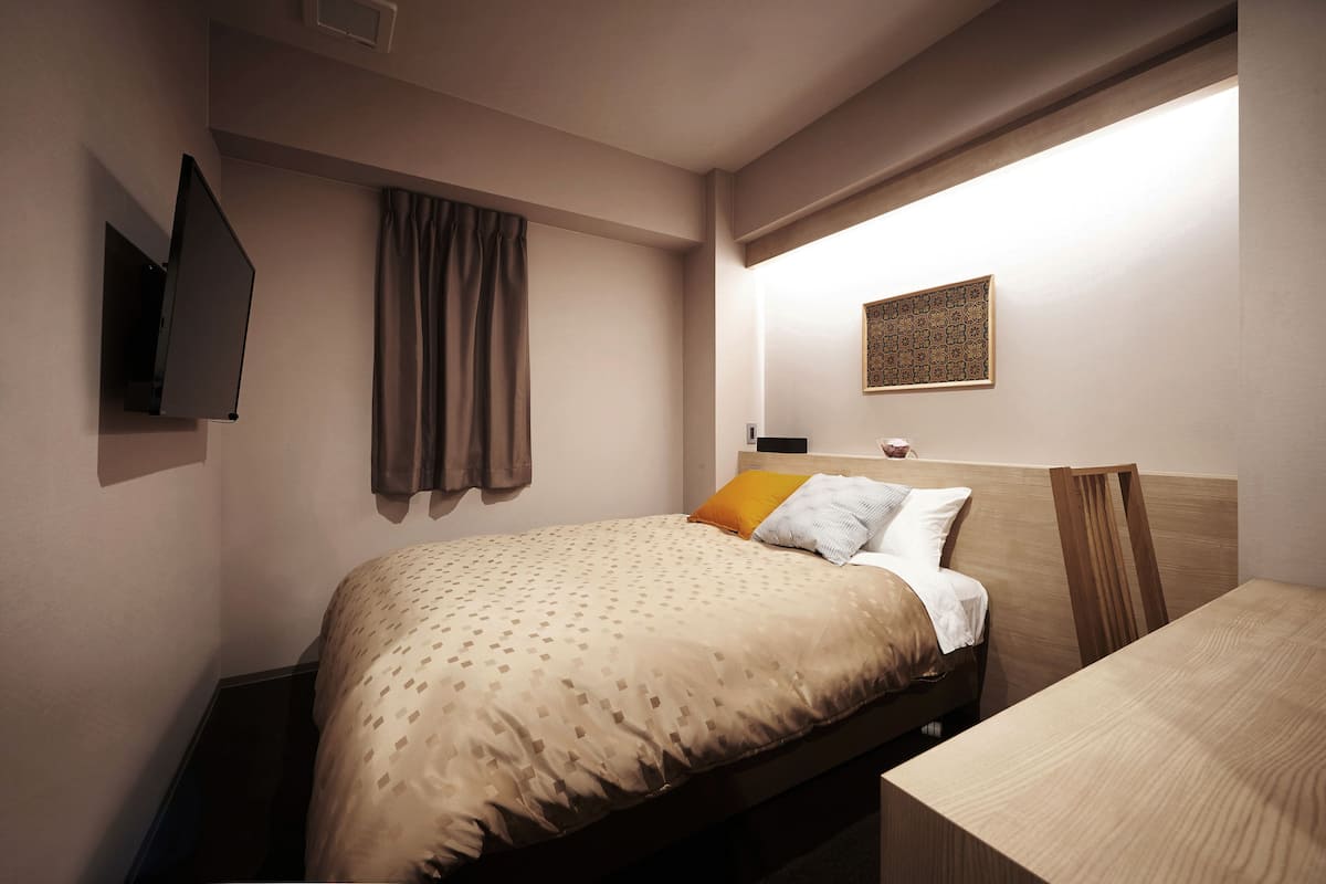 Habitación de hotel ordenada con cama, mesita de noche, televisor de pared, ventana con cortinas y un adorno en la pared.