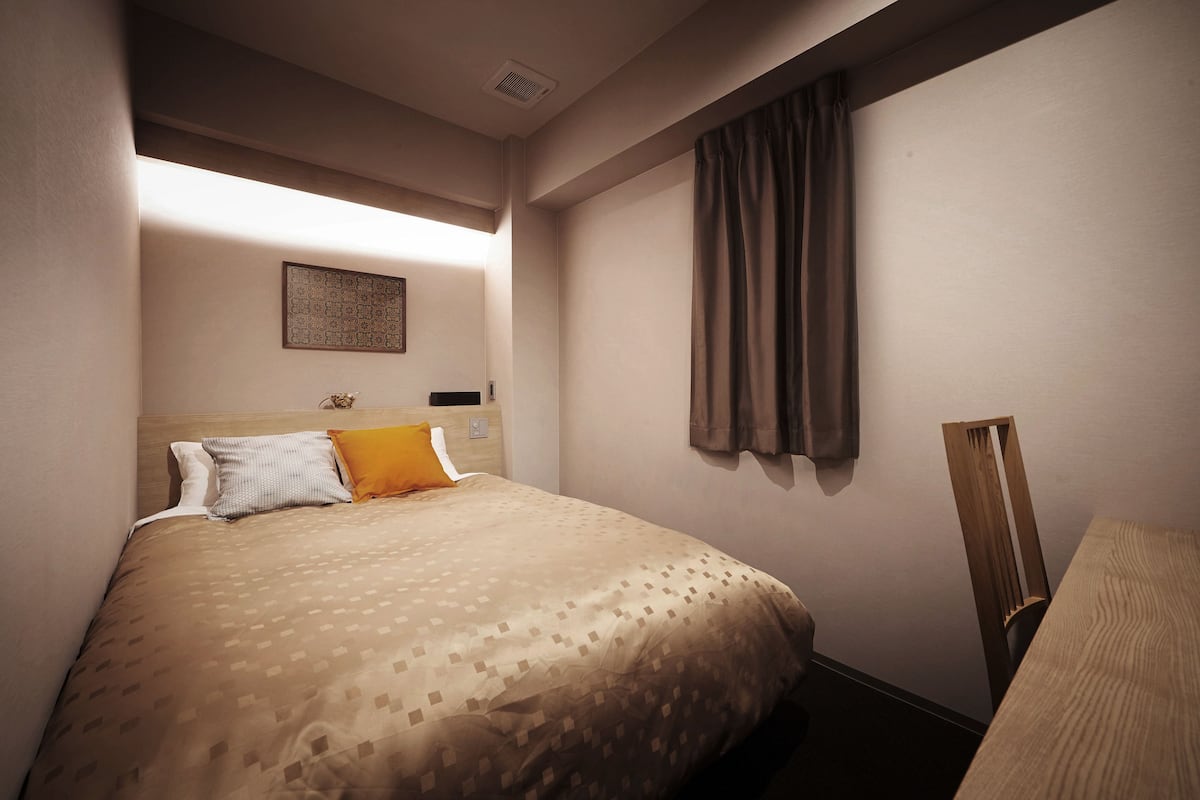 Habitación de hotel con una cama bien hecha, cabecera, una obra de arte en la pared, ventana con cortinas y una silla de madera.