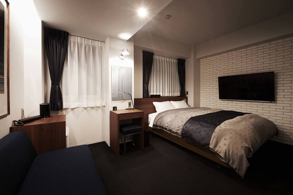 Una habitación de hotel con cama, escritorio, silla, televisor montado en la pared y ventana con cortinas.