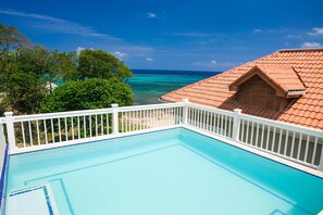 Une piscine d’un bleu éclatant, avec une terrasse et des garde-corps blancs, entourée d’une végétation luxuriante et offrant une vue sur l’océan.