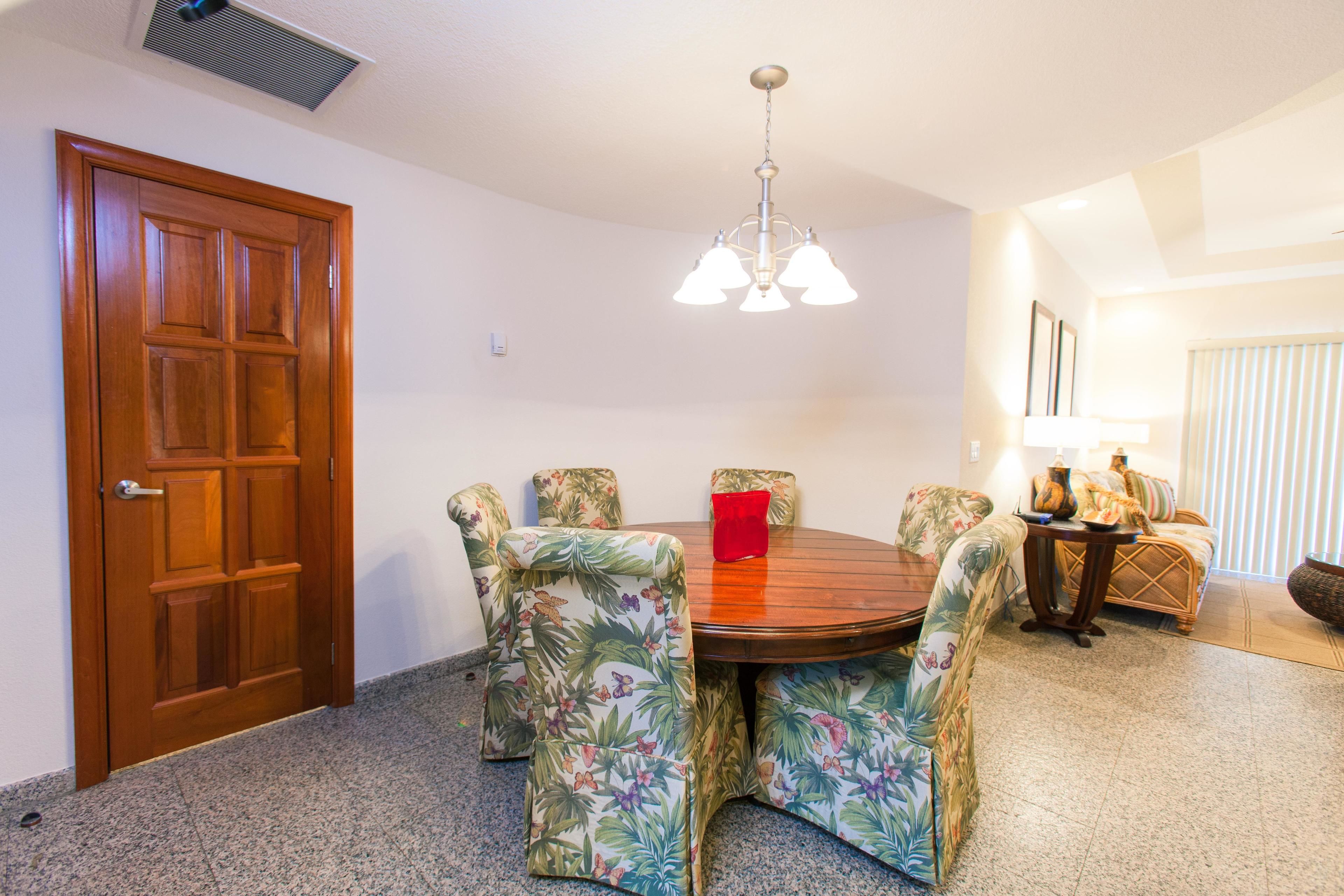A dining area with a wooden table, floral upholstered chairs, a red cushion, and a hanging light fixture.