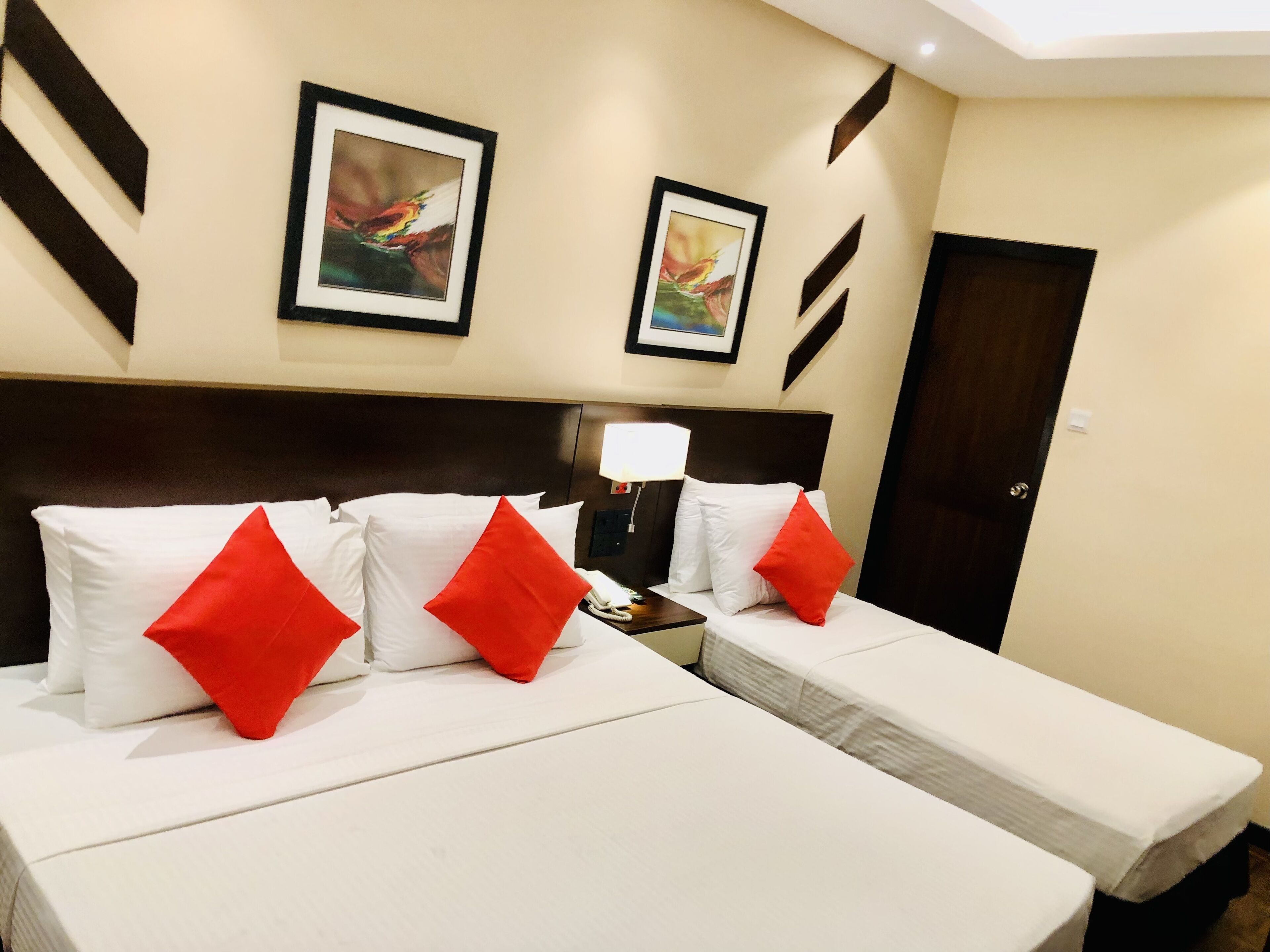 A hotel room with two beds, each with white bedding and red pillows. There are two framed pictures on the wall, a bedside lamp, and a door.