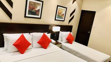 A hotel room with two beds, each with white bedding and red pillows. There are two framed pictures on the wall, a bedside lamp, and a door.