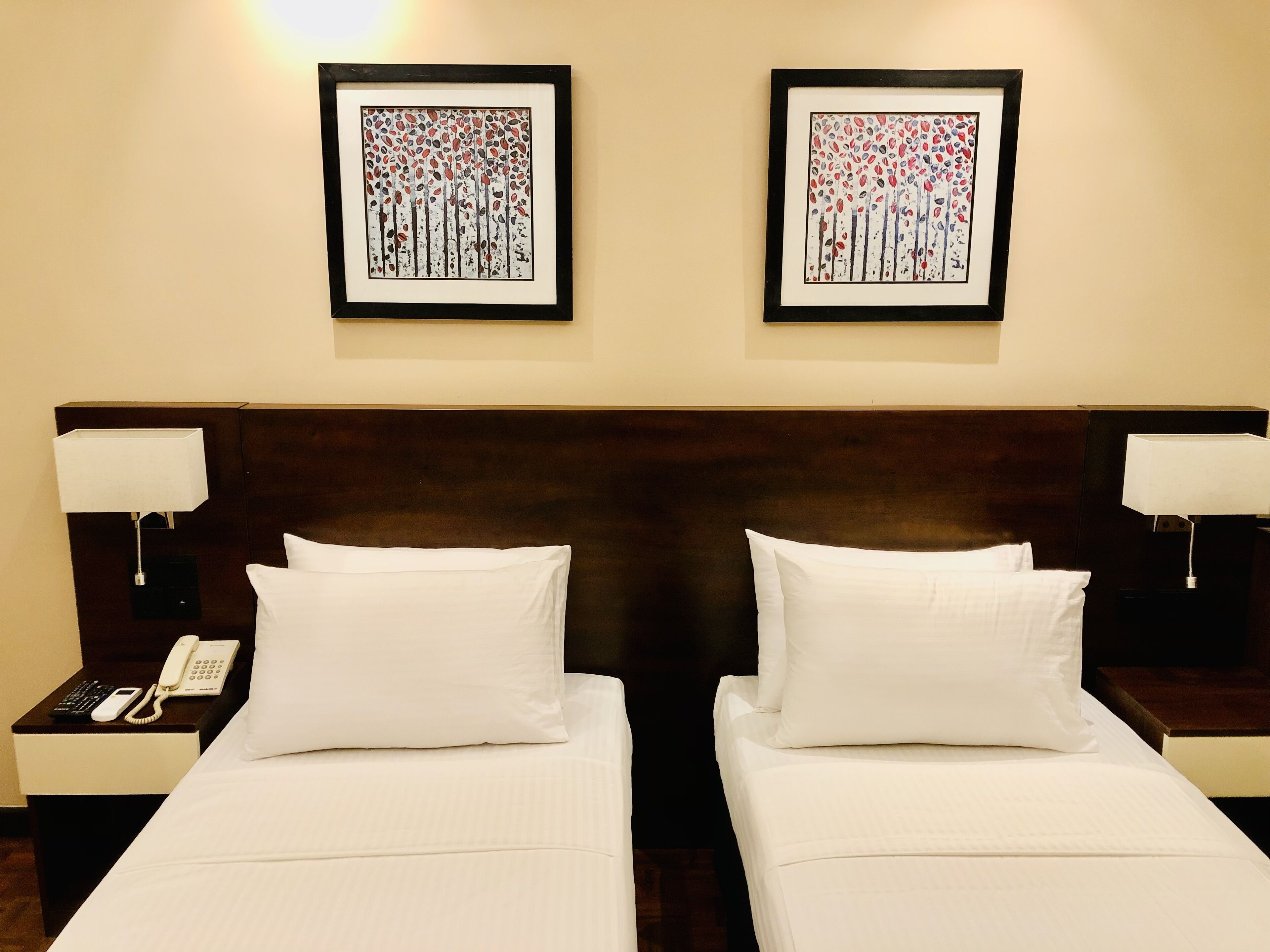 A hotel room with two beds, each with white bedding and pillows, a wooden headboard, bedside lamps, and two framed artworks on the wall.