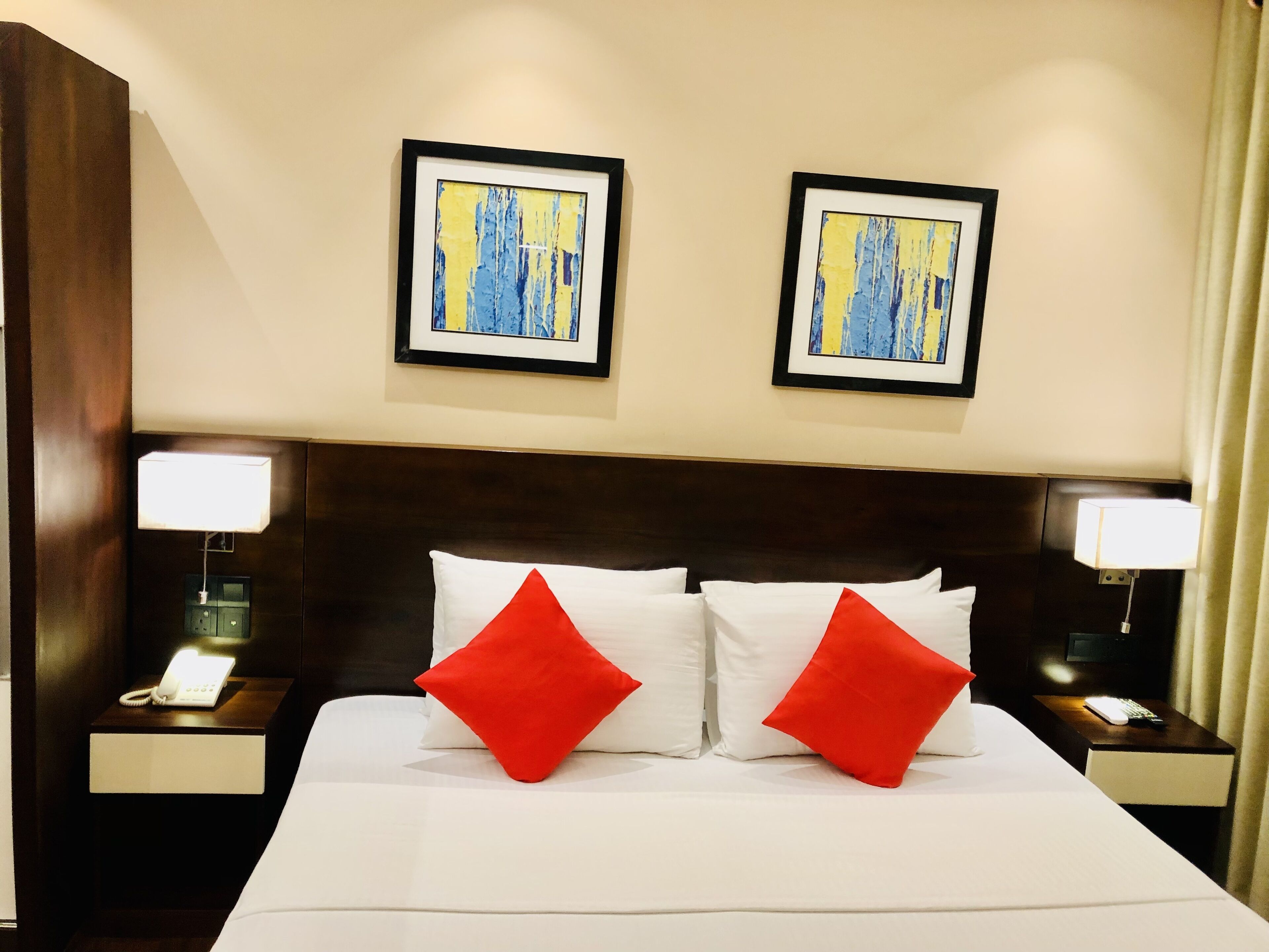 Habitación de hotel con una cama grande, dos mesitas de noche, un teléfono, dos almohadas rojas y dos cuadros abstractos enmarcados en la pared.