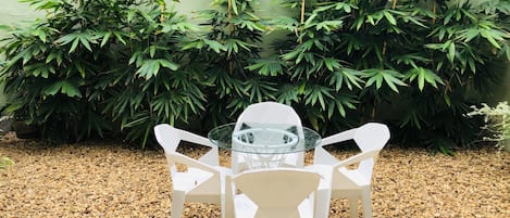 A table with four white chairs in a garden area with pebbles and green foliage in the background.