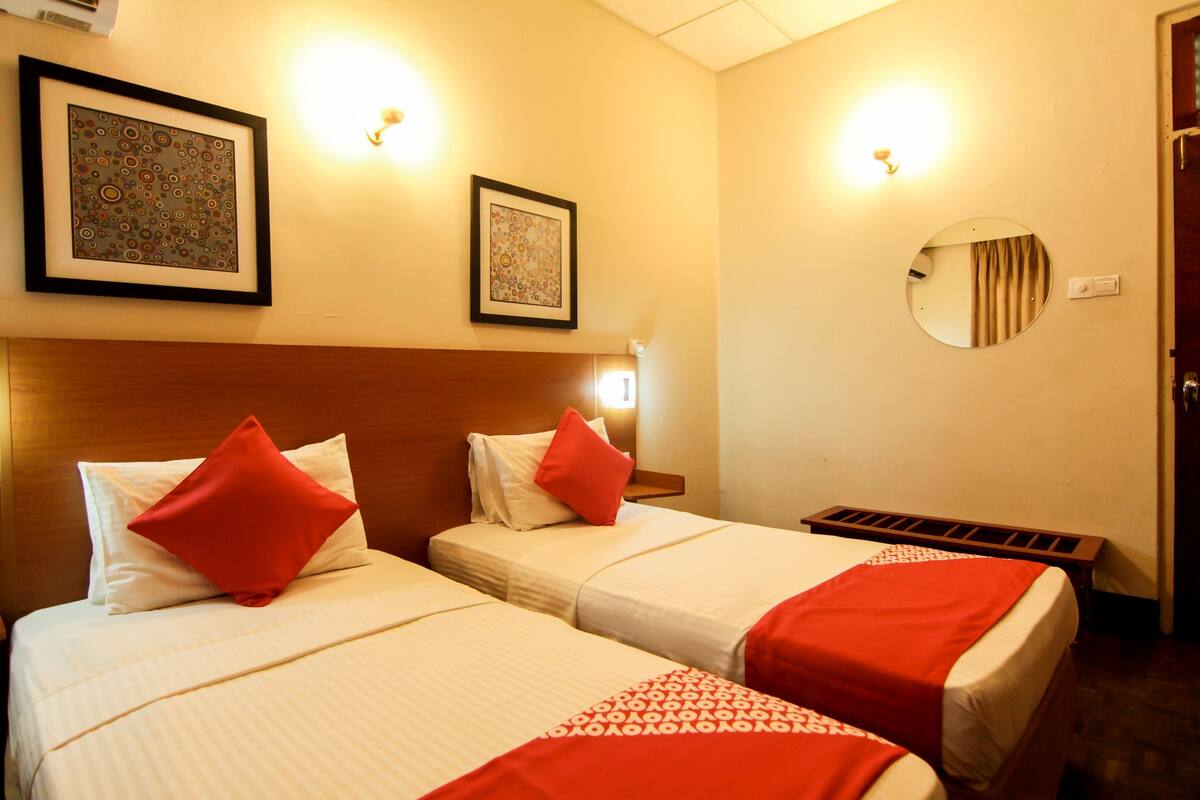 A hotel room with two beds, each with a red pillow, a headboard, a framed picture on the wall, and a round mirror.