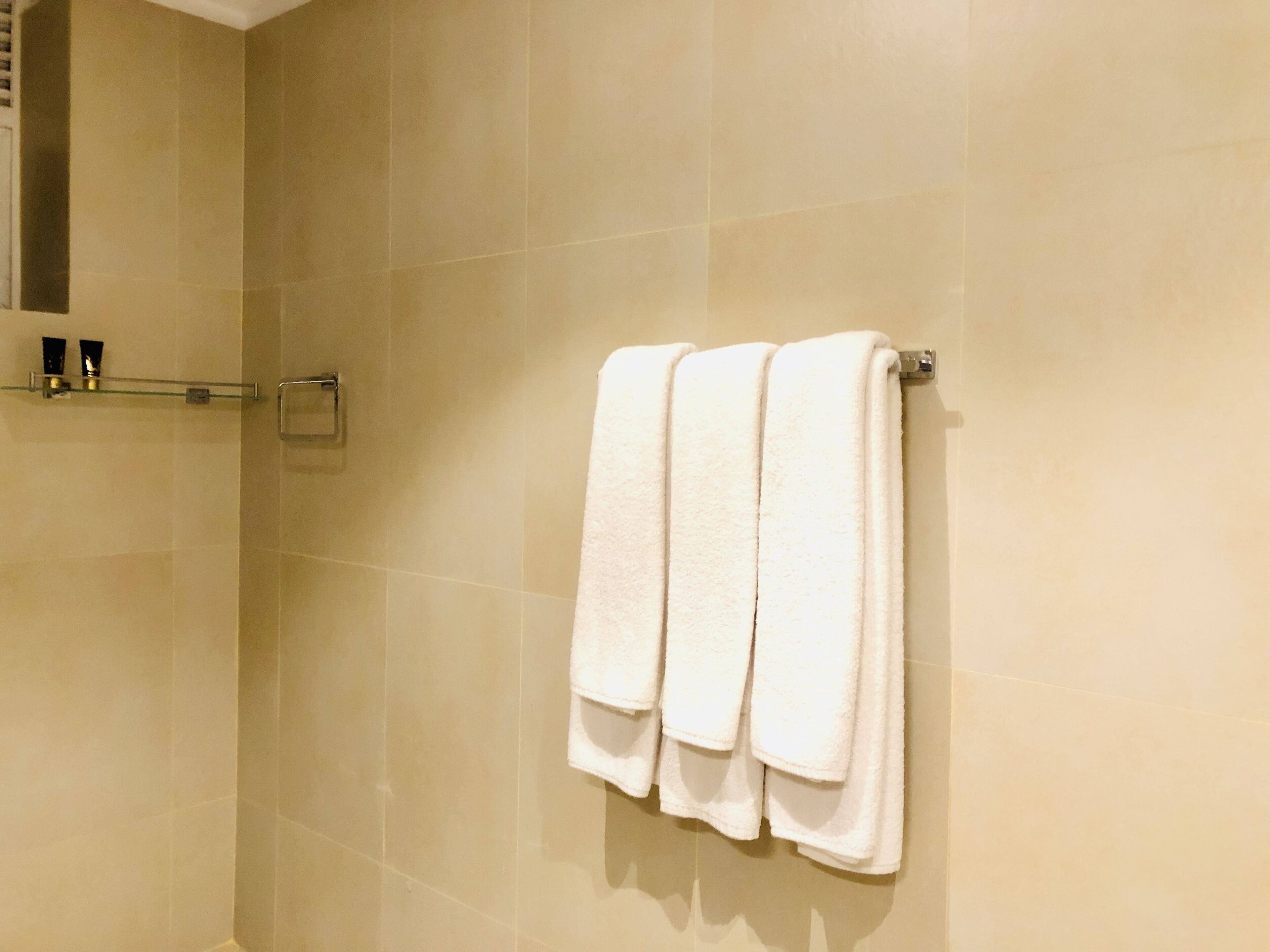 A bathroom with beige tiled walls, a glass shelf with toiletries, and a towel rack with three folded towels.