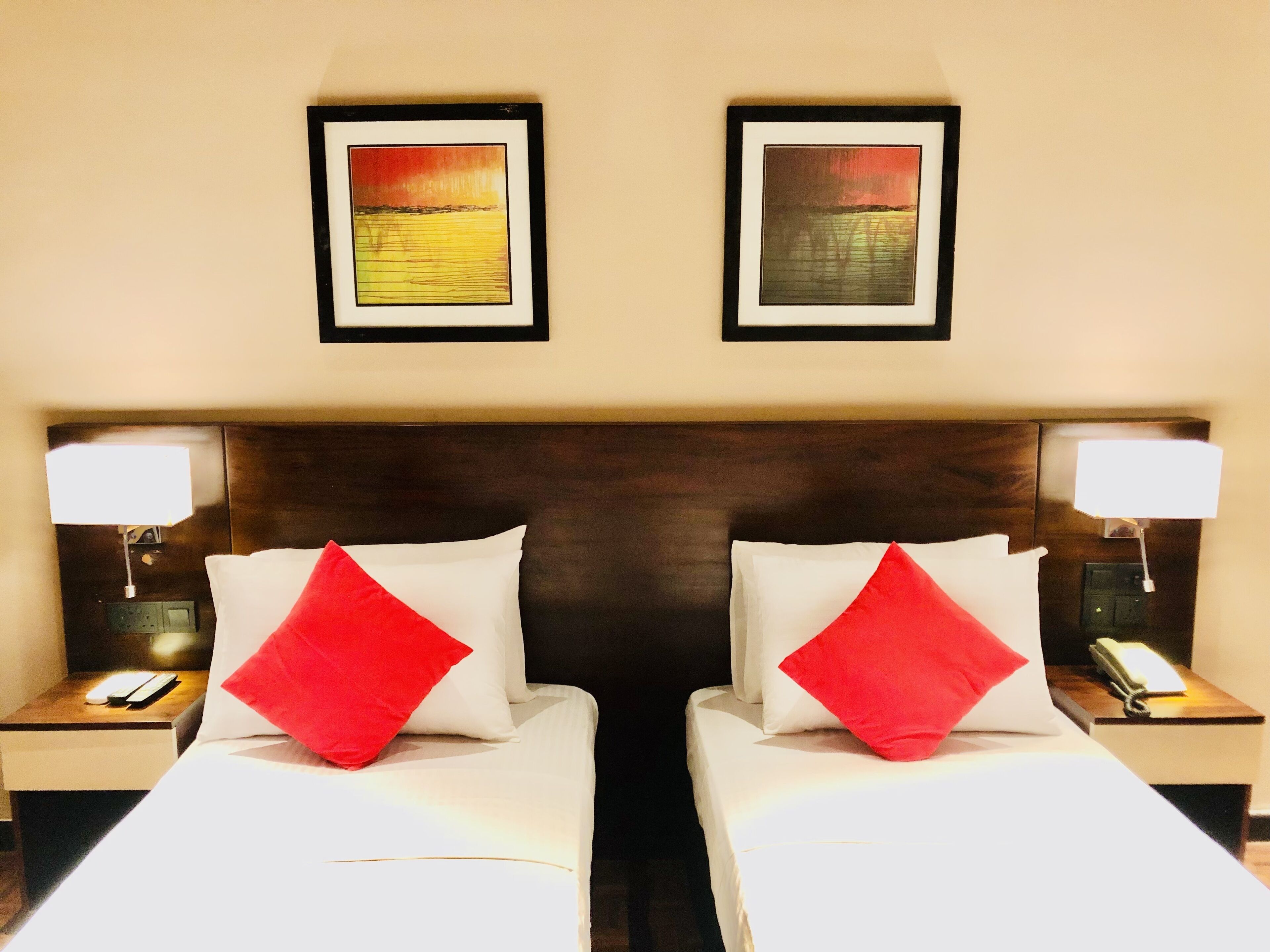 Una habitación de hotel con dos camas, cada una con una colcha blanca y un almohadón rojo. Hay cuadros enmarcados en la pared sobre las camas.