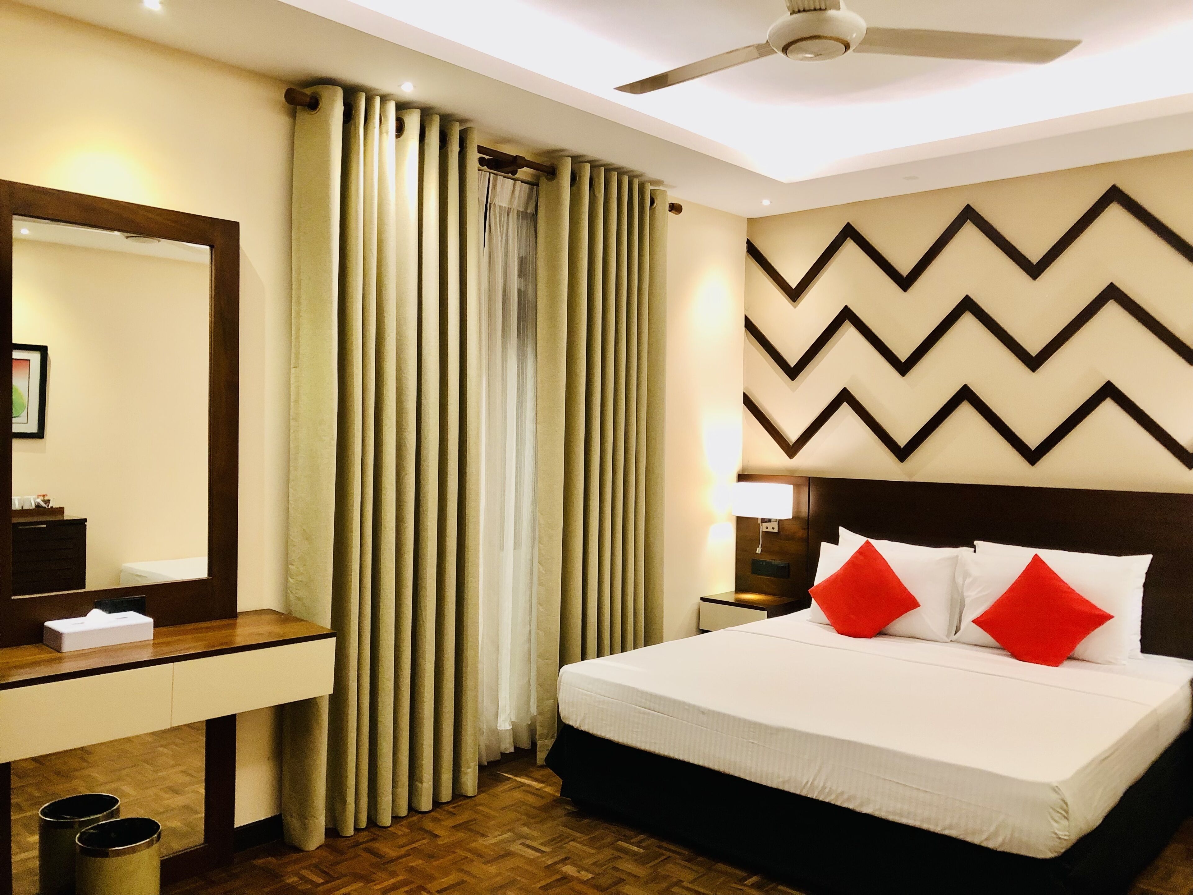 A hotel room with a bed, a nightstand, a mirror, a wooden dresser, and a window with curtains.