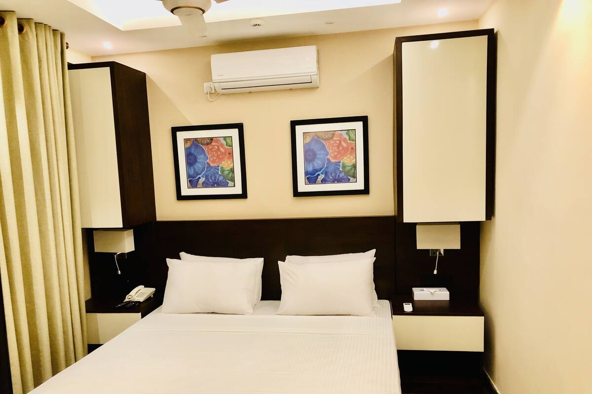 A hotel room with a large bed, two wall-mounted air conditioning units, and two framed floral paintings above the bed.
