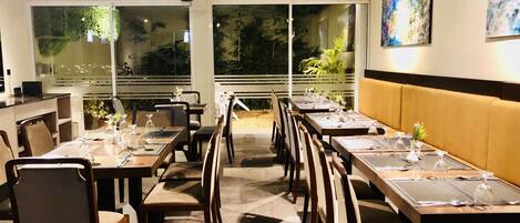 A restaurant with a long dining table, chairs, and a booth area. There are large windows with a view of greenery outside.