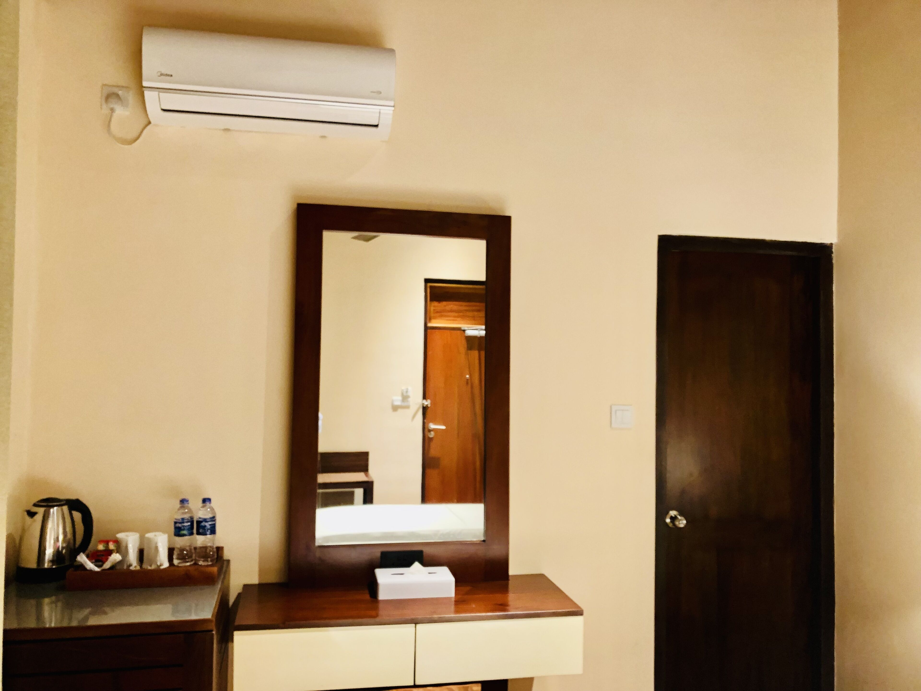 Une chambre d’hôtel avec une coiffeuse en bois, un miroir, une porte et un appareil de climatisation fixé au mur.