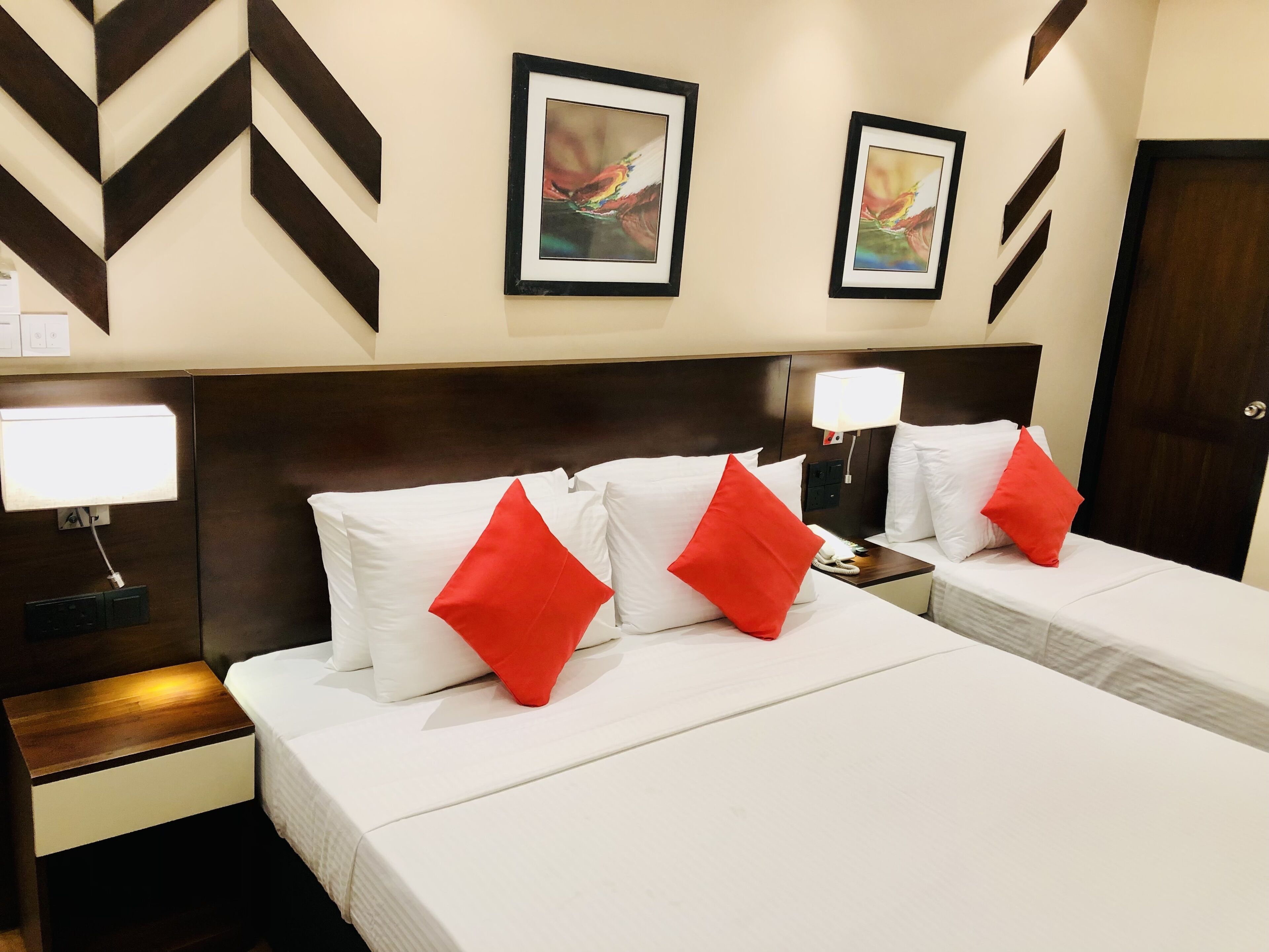 A hotel room with two beds, each with white bedding and red pillows. There are framed abstract paintings on the wall above the beds.