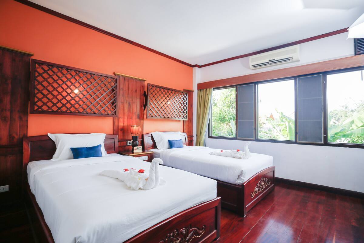 Une chambre d’hôtel avec deux lits, des têtes de lit en bois, une fenêtre donnant sur un paysage verdoyant et un appareil de climatisation.