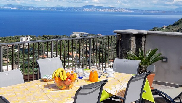 Une table dressée avec des fruits et des boissons sur un balcon donnant sur une vue de la côte.