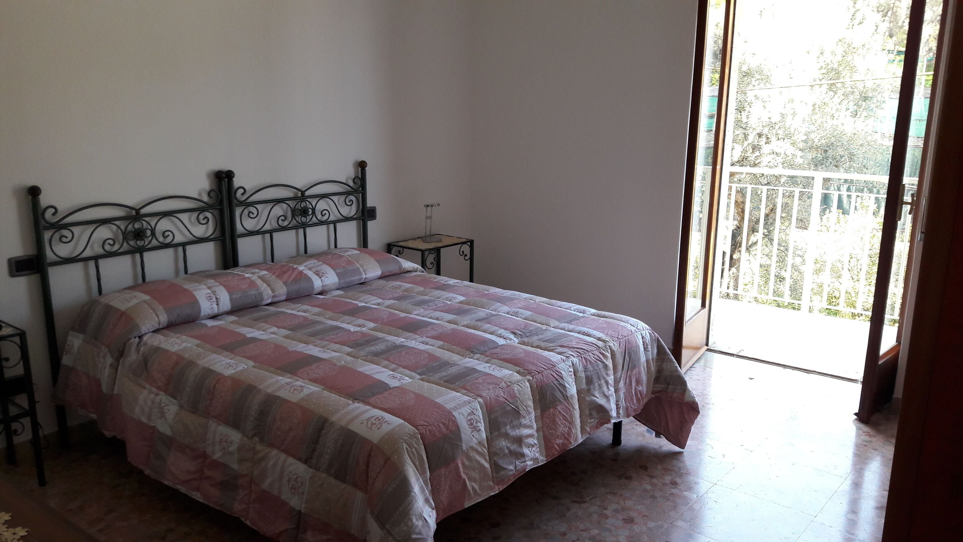 A bedroom with a bed, a nightstand, a window with a view of greenery, and a balcony.