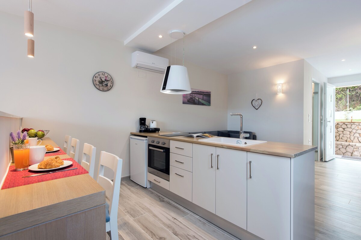 A modern kitchen with white cabinetry, a built-in oven, and a sink. There is a dining table with chairs and a breakfast setting.