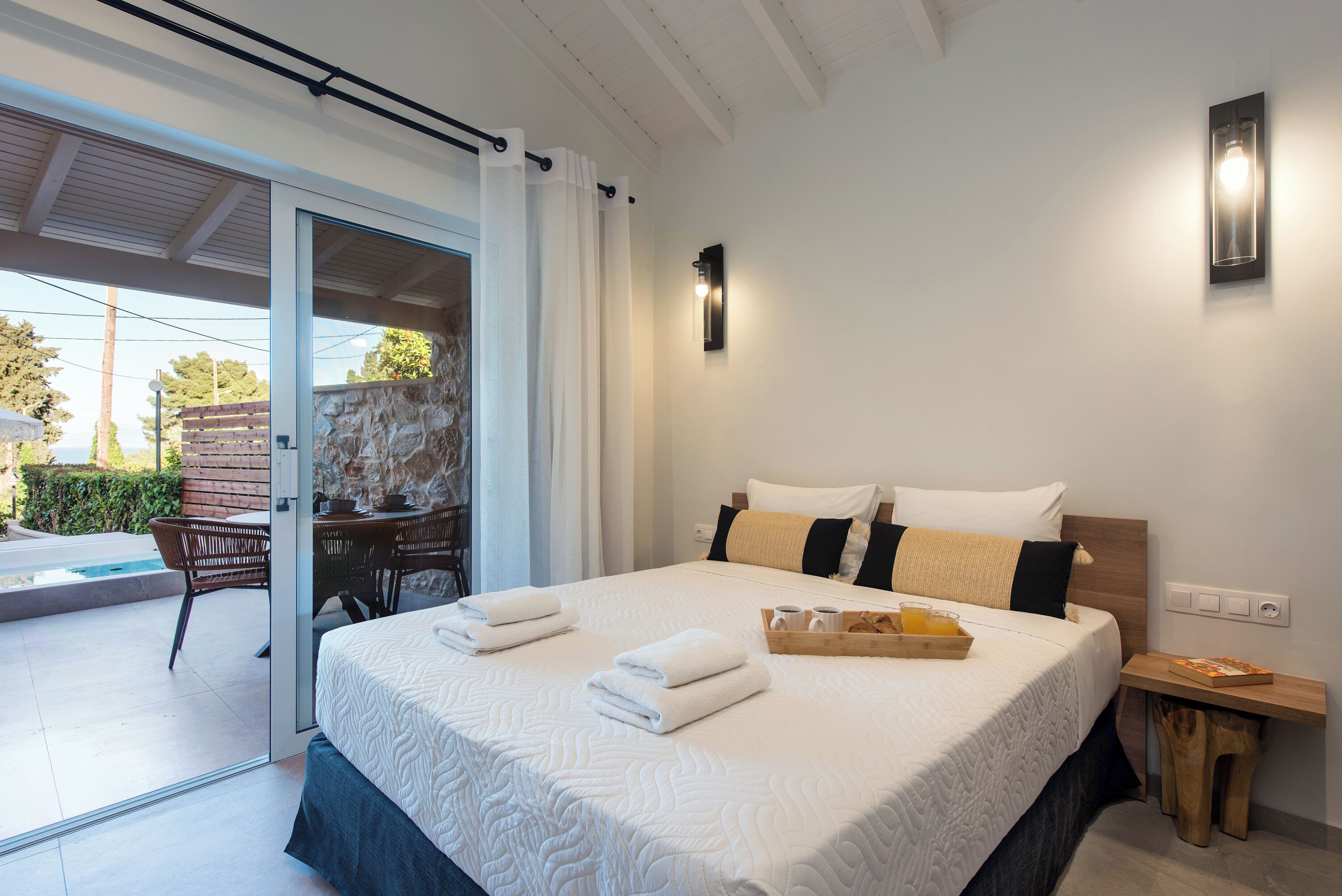 A modern hotel room with a large bed, a small table with a tray of breakfast items, a wall-mounted light, and a view of the outdoor area.