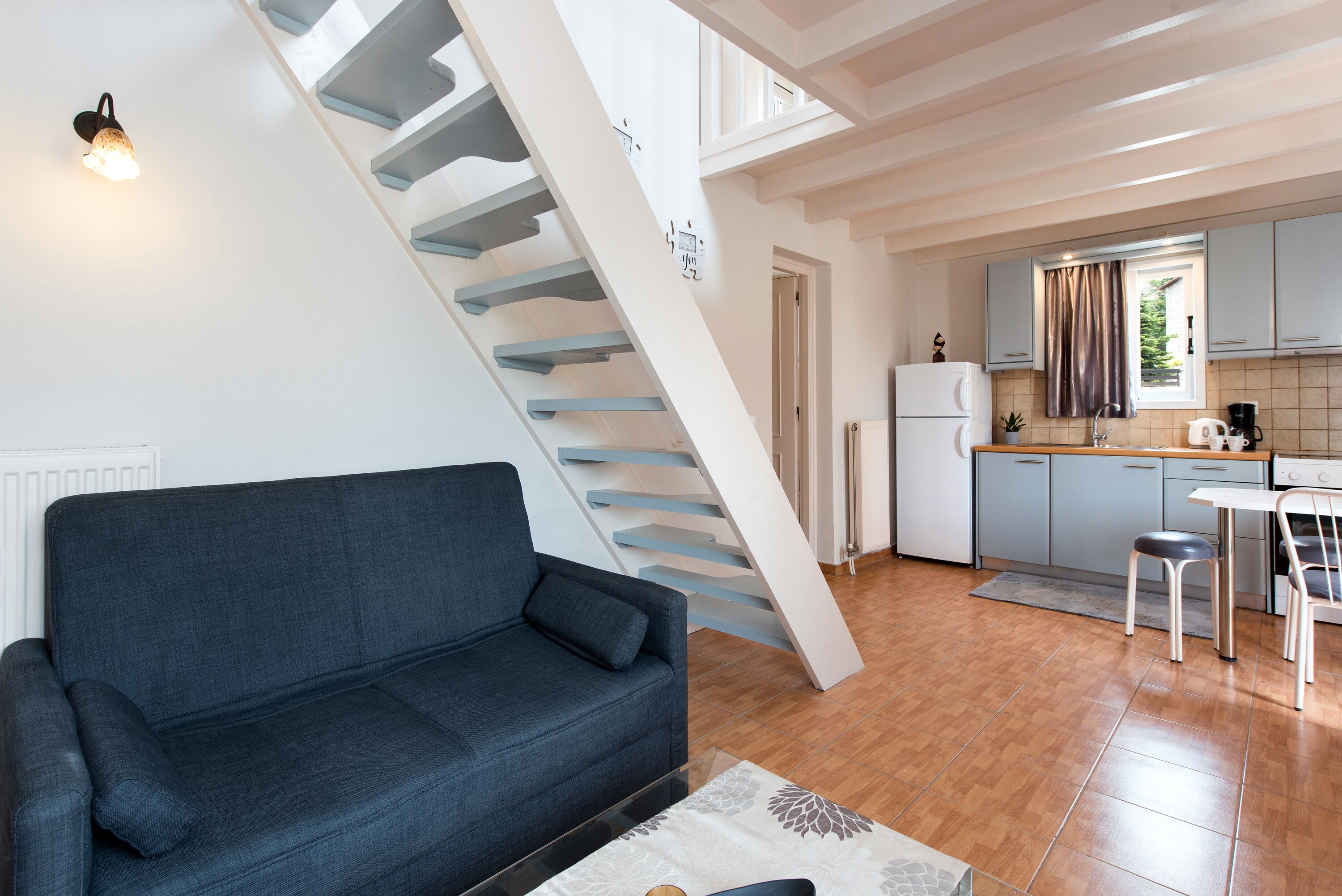 A modern living room with a dark blue sofa, a staircase with white railings, a kitchen area with light-colored cabinets, and a dining table with two chairs.