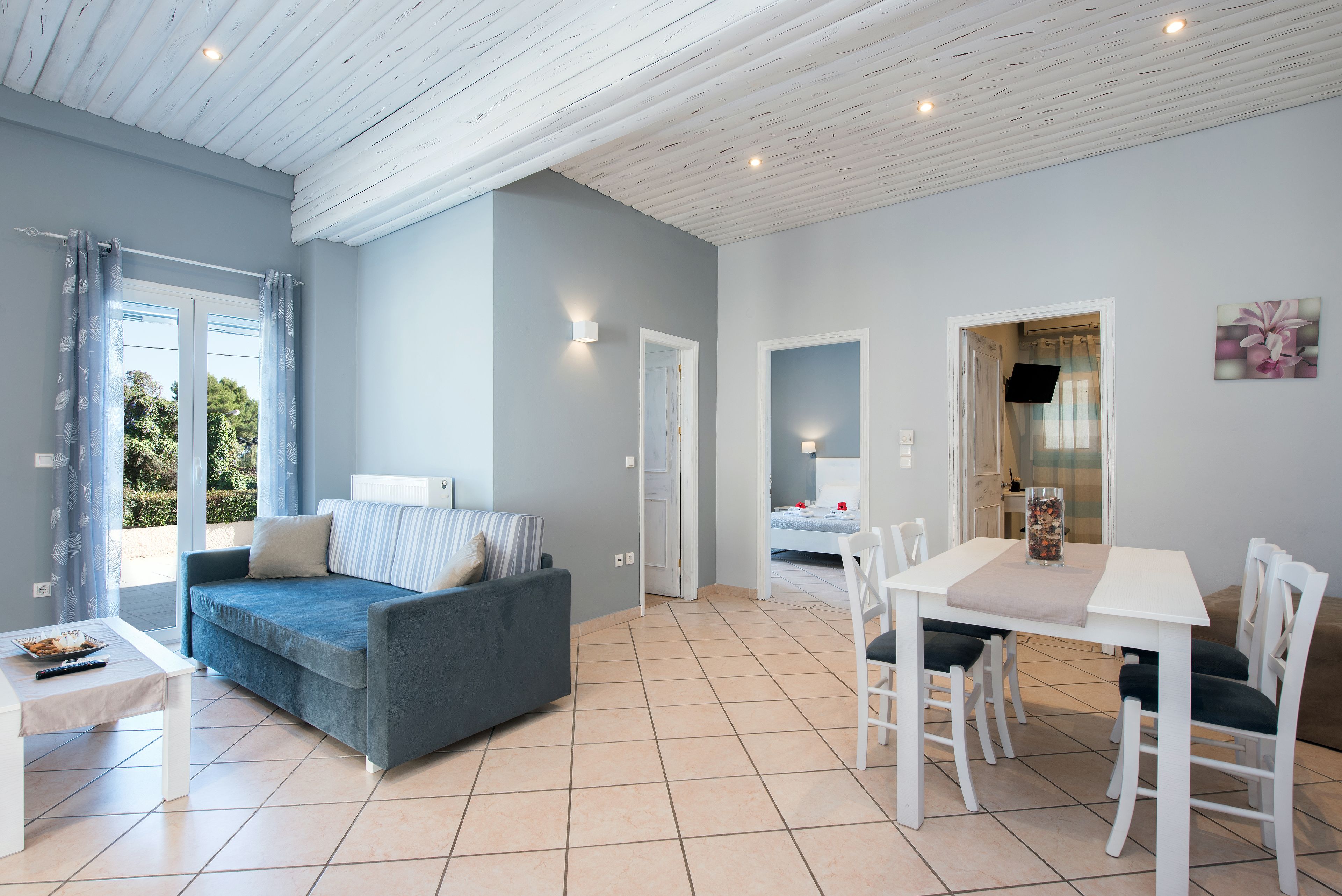 Una sala de estar moderna con un sofá gris, una mesa de comedor blanca con sillas, y una cocina visible a través de una puerta abierta.