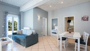 Una sala de estar moderna con un sofá gris, una mesa de comedor blanca con sillas, y una cocina visible a través de una puerta abierta.