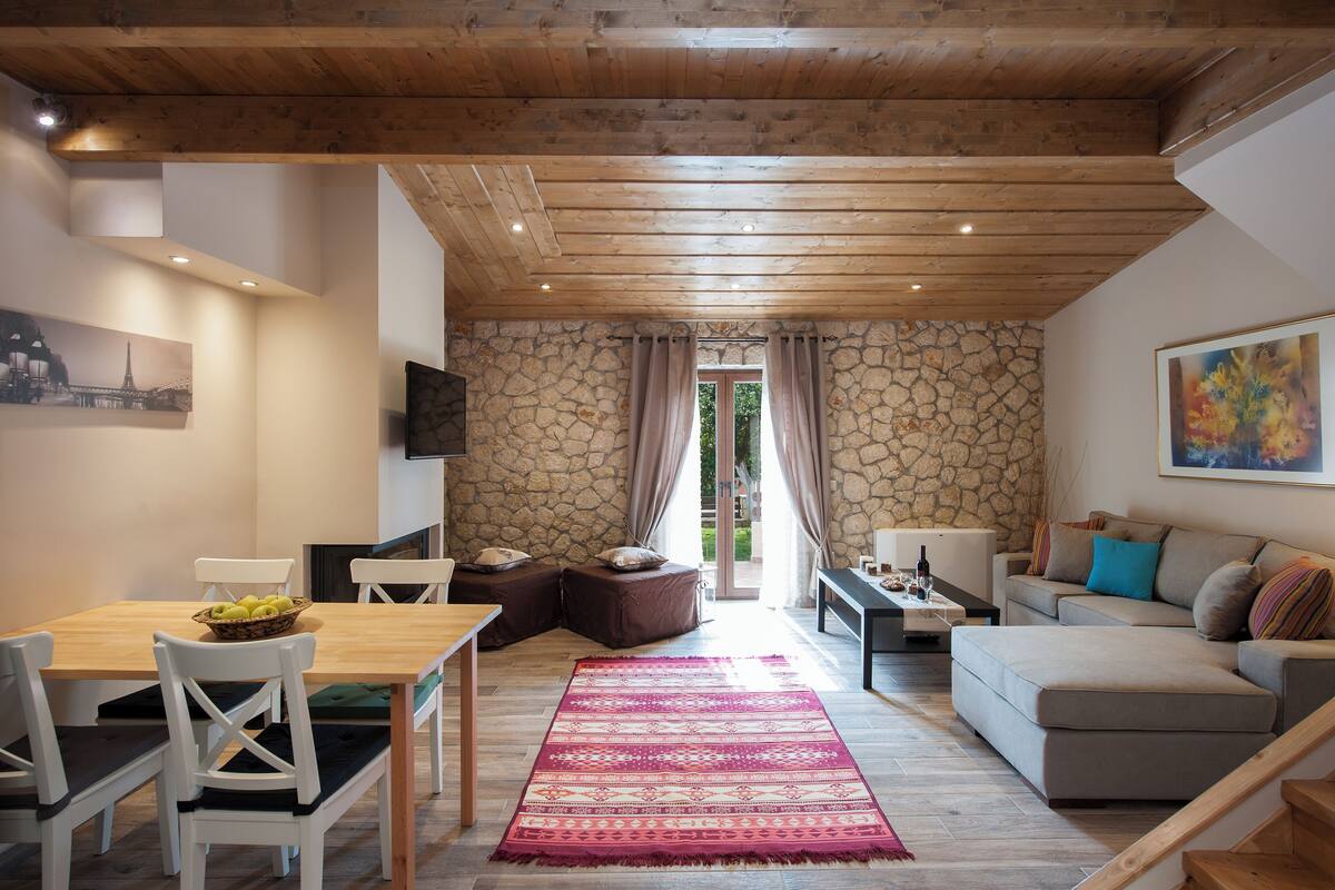 A dining area with a wooden table and chairs, a stone wall, a sofa, and a colorful rug.