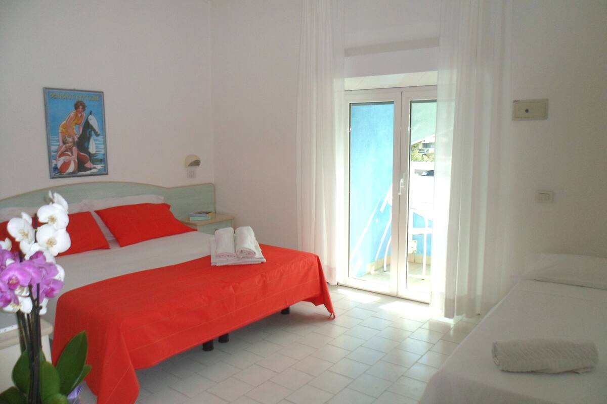 A hotel room with a bed, a nightstand, a window with curtains, and a view of the outside.