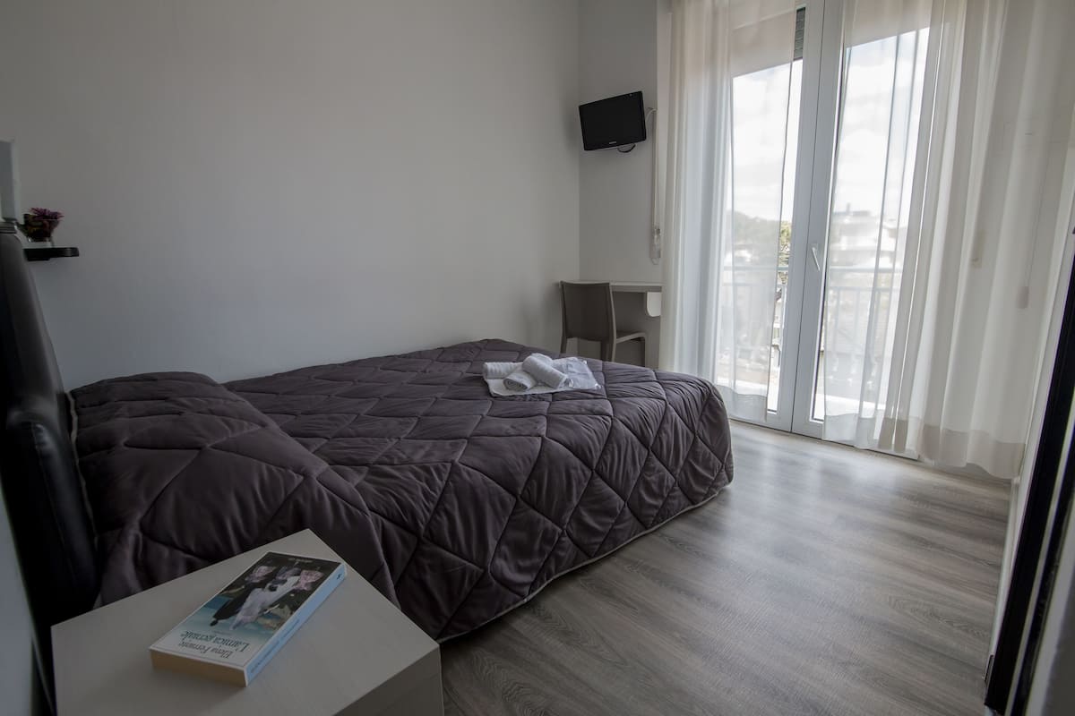 A bedroom with a bed, a bedside table with a book, a chair, a TV on the wall, and a sliding glass door to the outside.