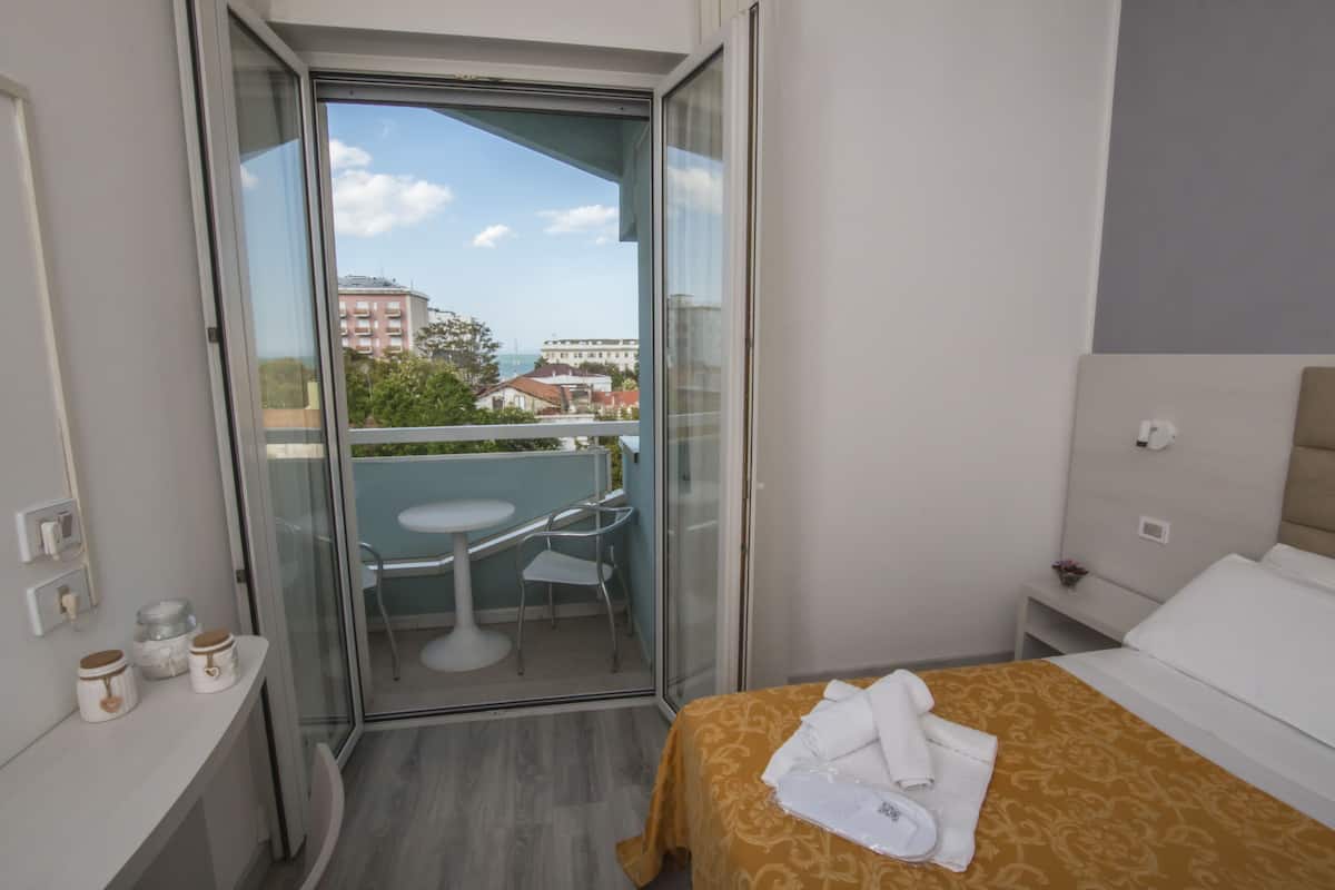 A hotel room with a bed, a small table, a chair, and a glass door leading to a balcony.