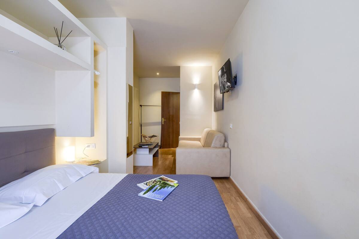 A hotel room with a bed, a nightstand, a desk, a chair, and a TV mounted on the wall.