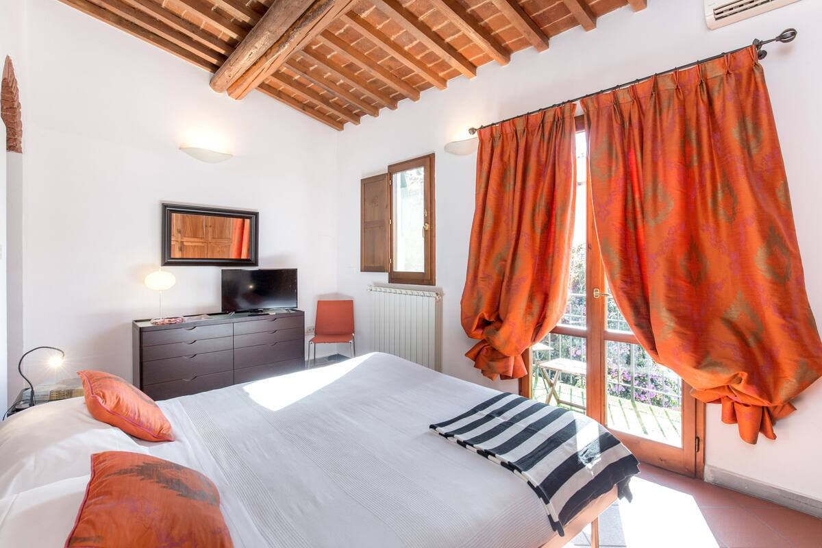 A bedroom with a bed, a dresser, a TV, a window with curtains, and a radiator.