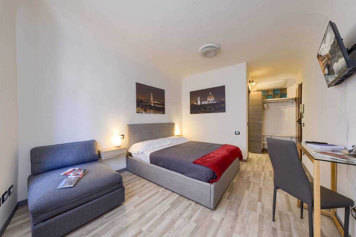 A modern hotel room with a bed, a desk, a chair, and two wall-mounted TVs.