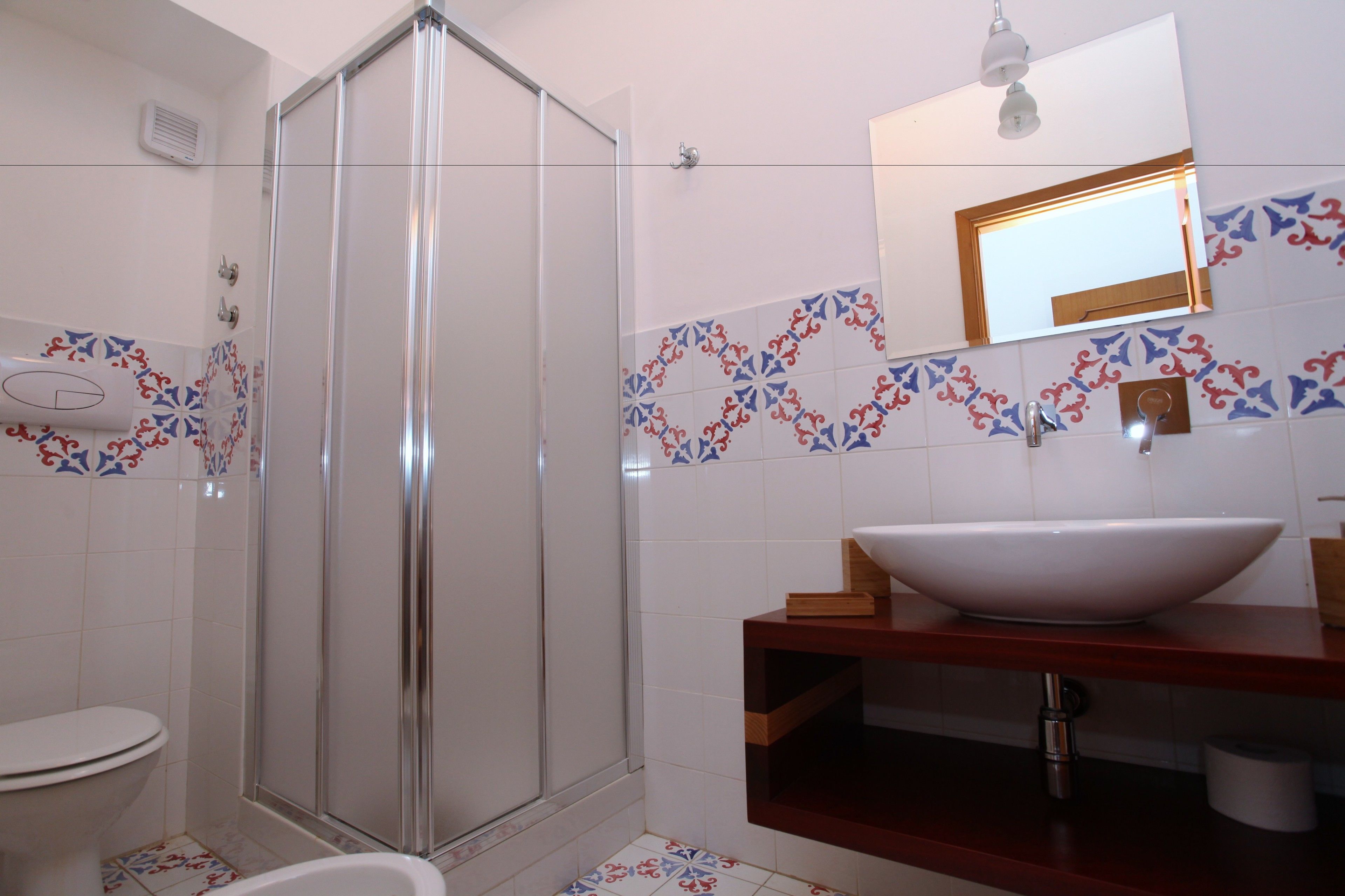 Ein Badezimmer mit Duschkabine aus Glas, einem weißen Waschbecken mit Holzablage und Wandfliesen mit Muster.