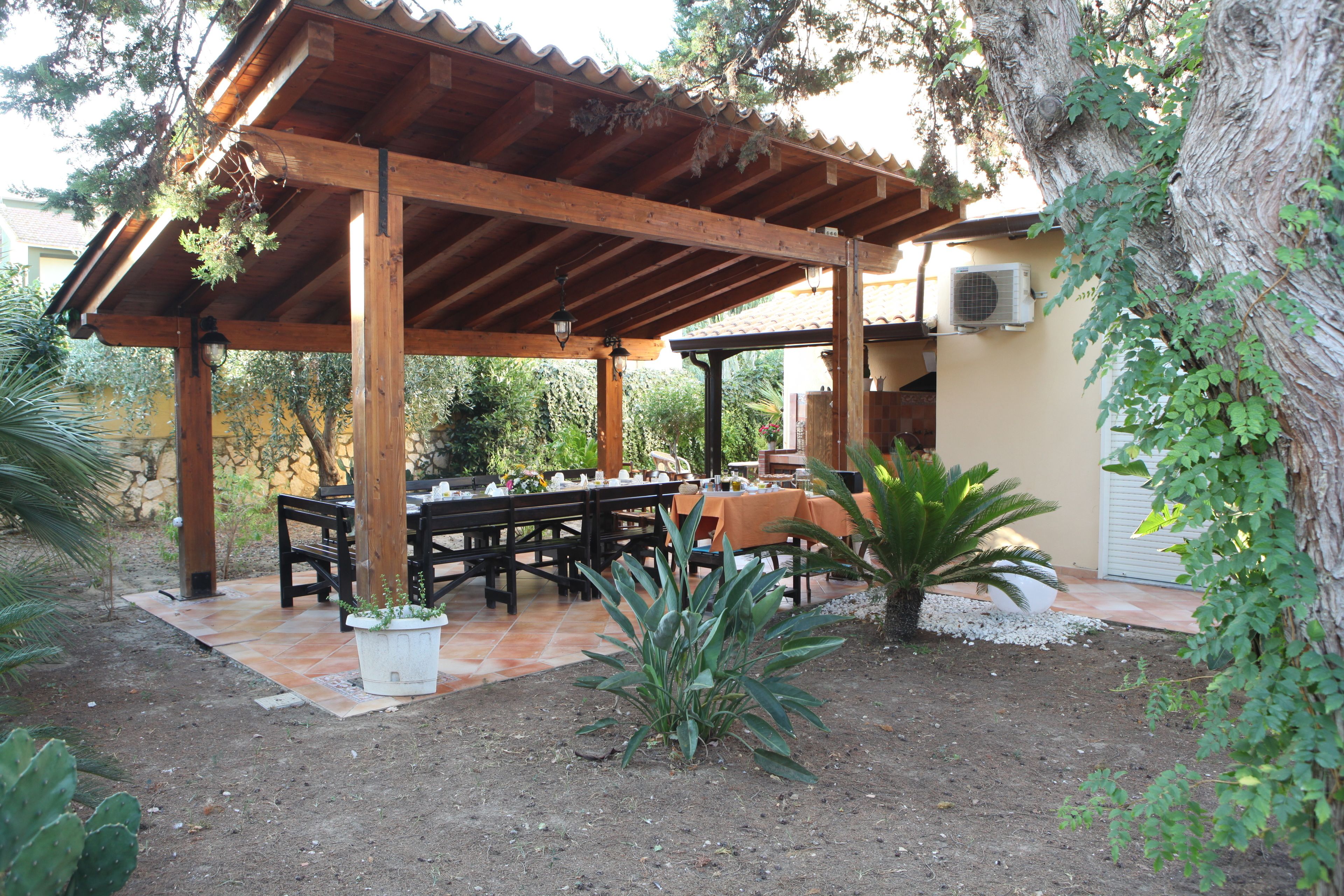 Un espacio de comedor al aire libre cubierto, con mesas y bancos de madera, rodeado de plantas en macetas y árboles.