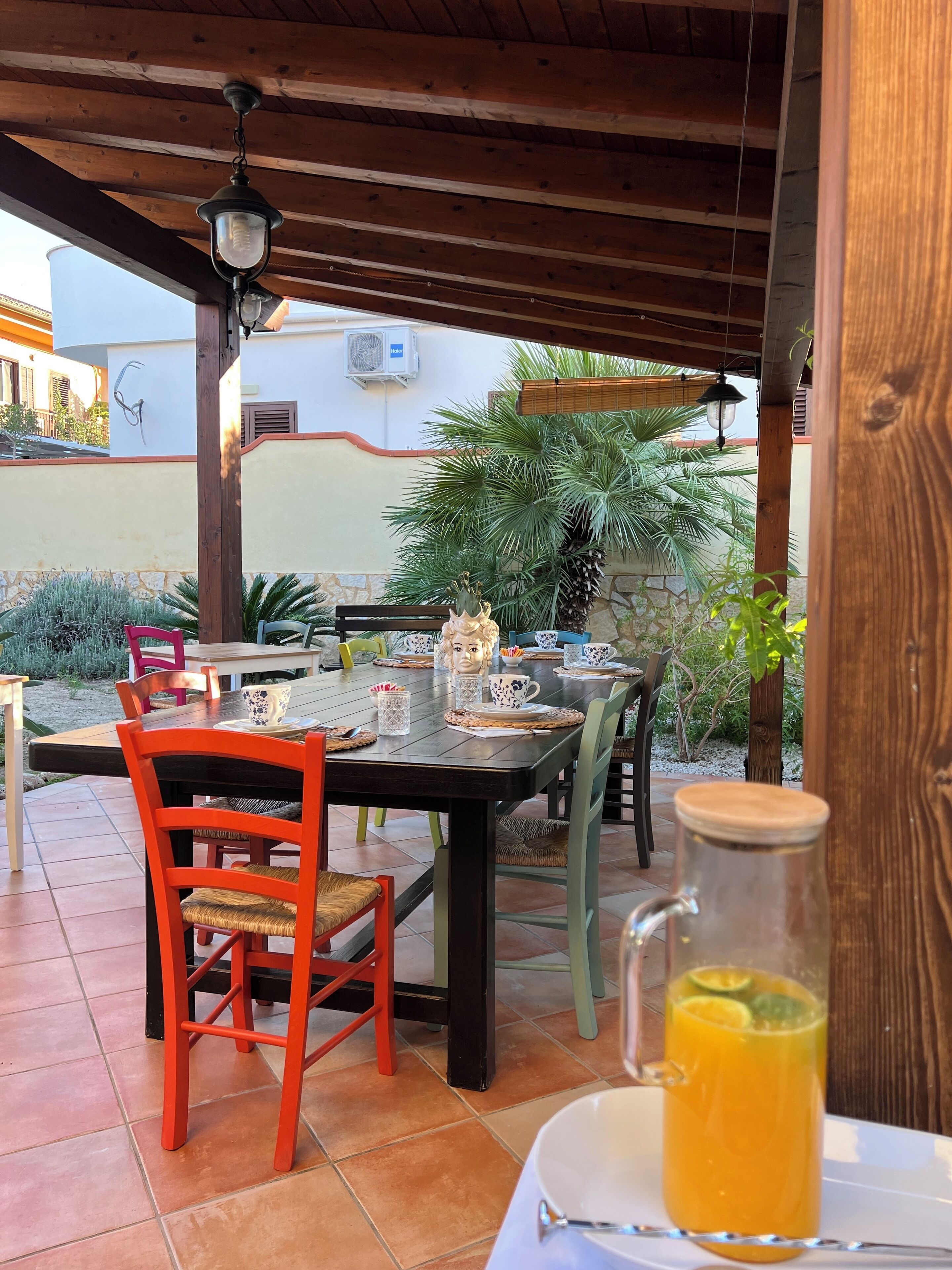 A covered outdoor dining area with a table set for two, surrounded by chairs, and a pitcher of orange juice on a table.