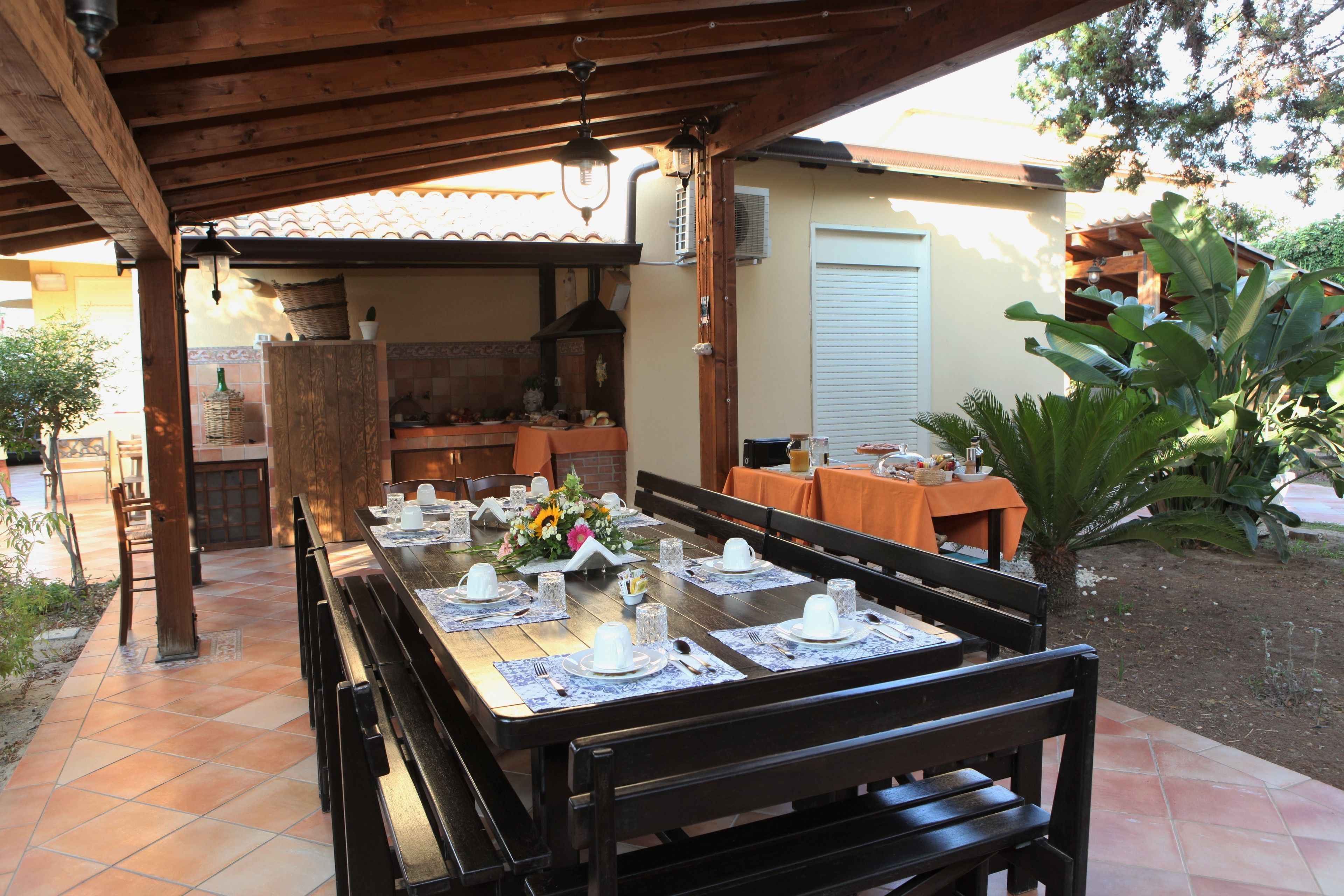 An outdoor dining area with a wooden pergola, a brick oven, and a dining table set for a meal.