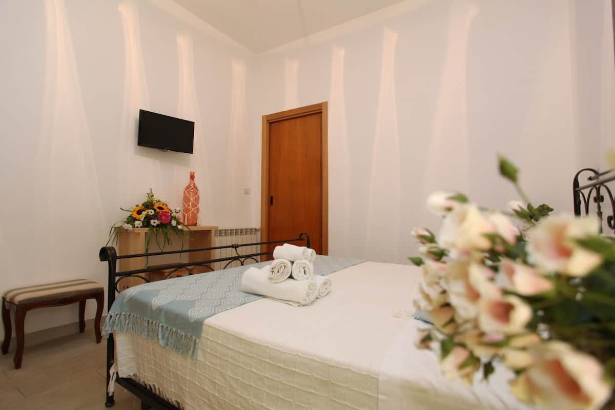 Une chambre à coucher avec un lit, une table de chevet ornée d’un vase de fleurs, un banc en bois et un téléviseur fixé au mur.