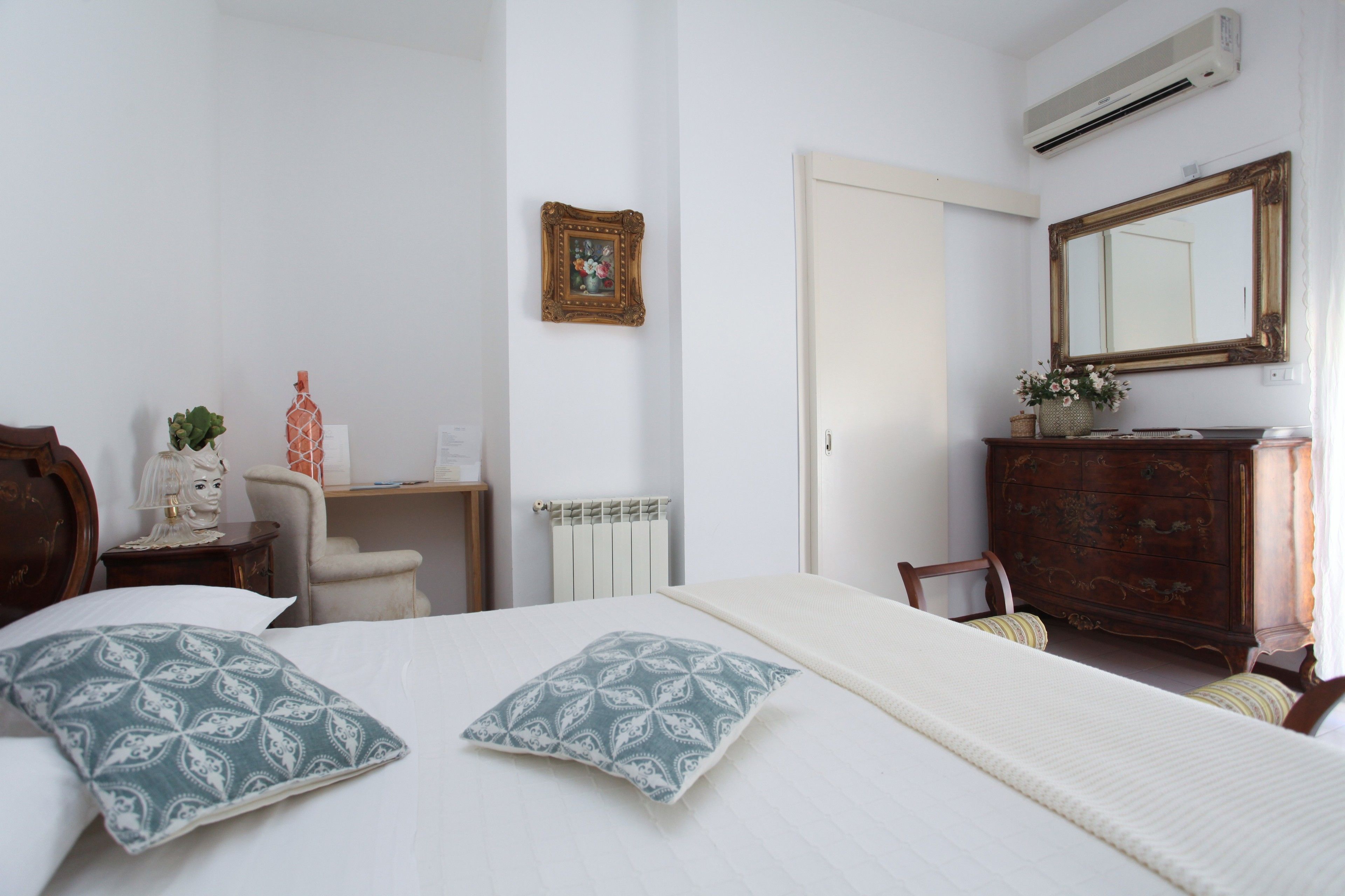 A bedroom with a bed, a dresser, a chair, a desk, a radiator, a picture frame, and a mirror.