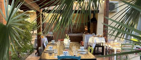 An outdoor dining area with wooden tables and chairs, surrounded by plants and a pergola.