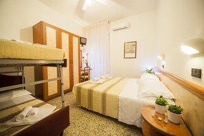 A hotel room with a bunk bed, a single bed, a wooden headboard, a small table with two potted plants, and a wall-mounted air conditioner. - Hotel Lily (Rimini)