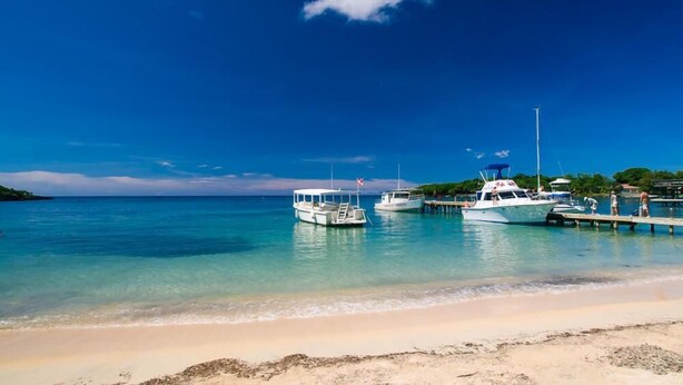 A beach with clear turquoise water, boats docked at a pier, and a sandy shore. - Sunset Villas 12I (Roatan)