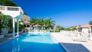 A swimming pool with white railings, palm trees, and buildings with balconies. - Sunset Villas 12I (Roatan)