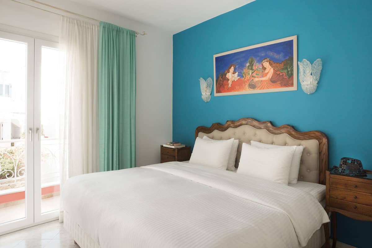 A bedroom with a large bed, a nightstand, a painting, and a view of the outdoors.